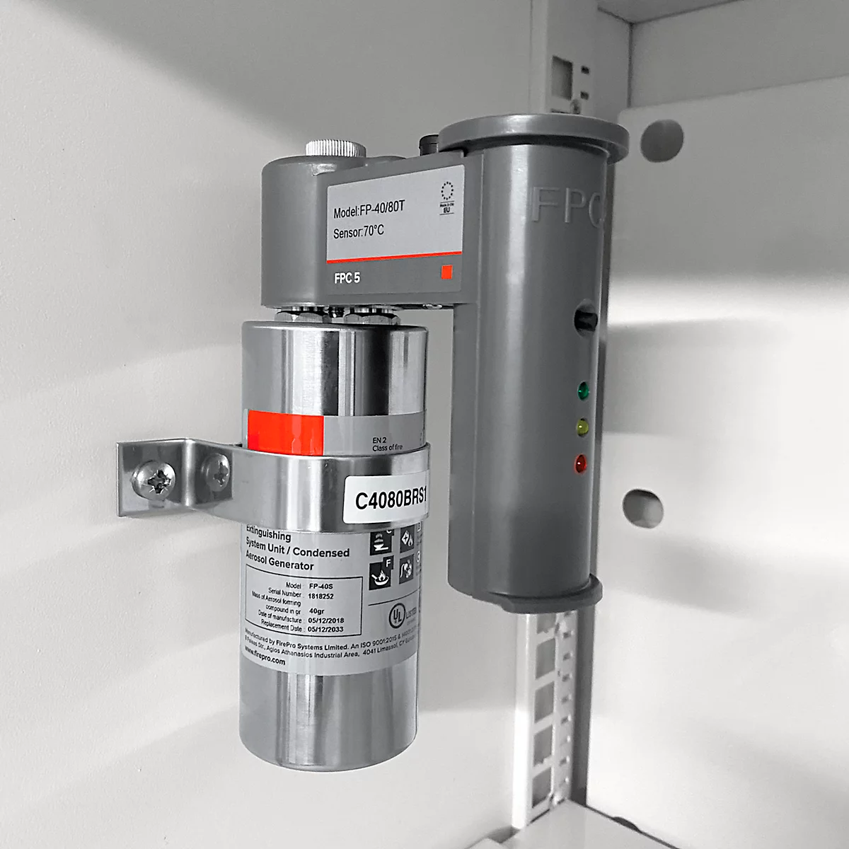 Grauer Brandschutzsensor mit roten und grünen LEDs. Der Sensor ist an einer weißen Wand in einem Schrank montiert.