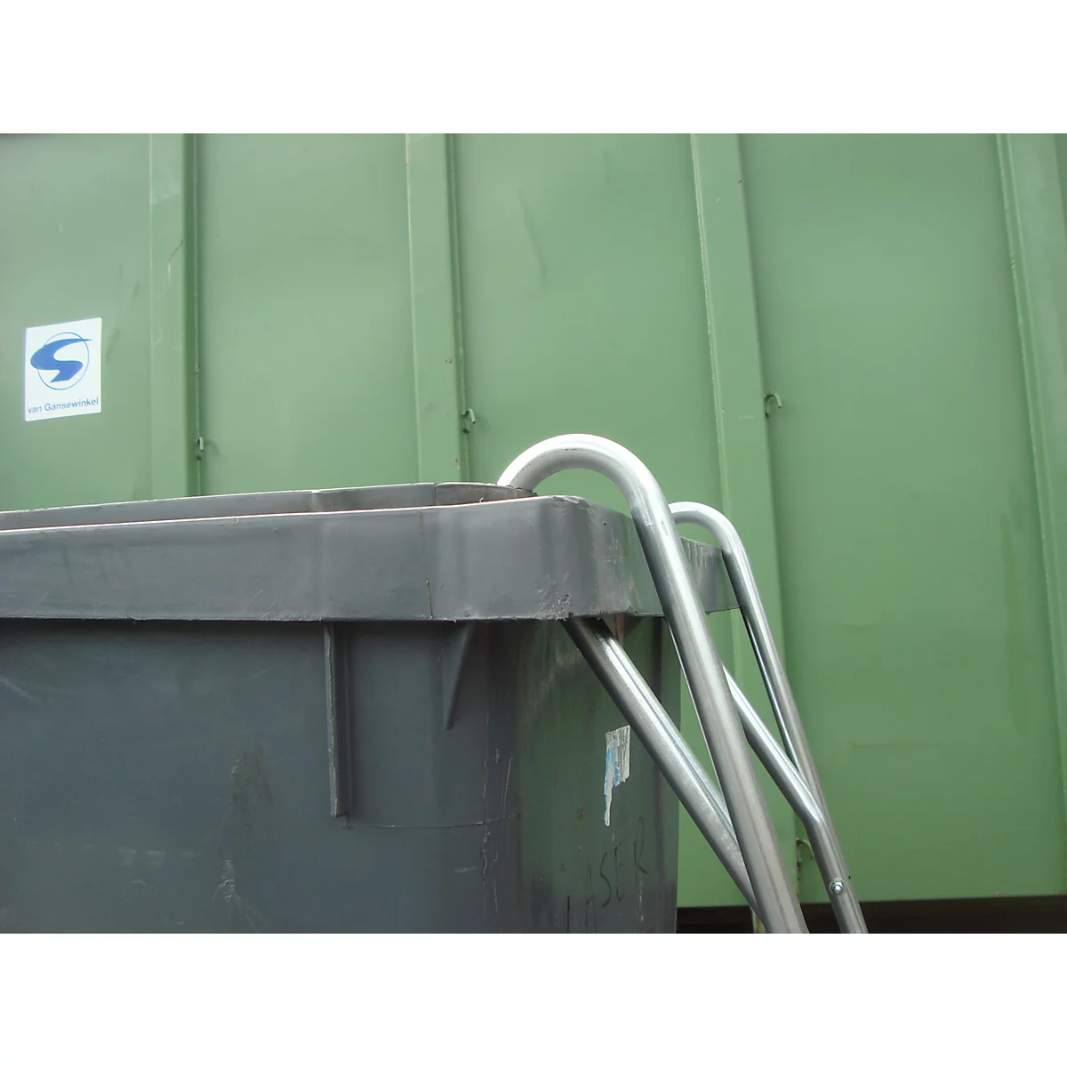 Nahaufnahme eines grauen Müllcontainers mit Metallgriff. Im Hintergrund eine grüne Wand.