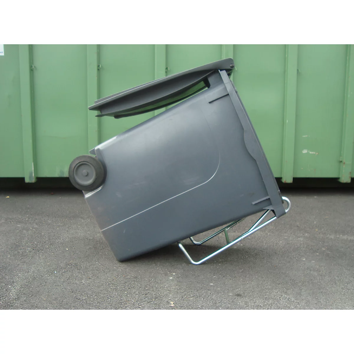 Ein grauer Mülleimer kippt auf einer befestigten Fläche. Im Hintergrund ein grüner Container.