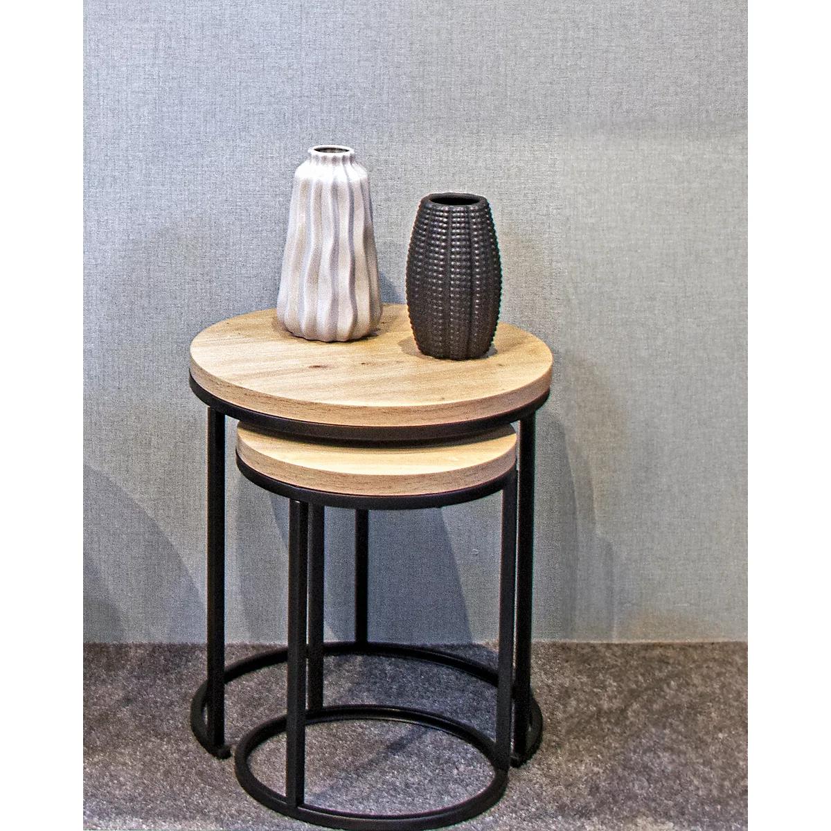 Zwei runde Beistelltische mit Vasen in einem Wohnraum. Oben Holz, schwarze Metallbeine, grauer Hintergrund.