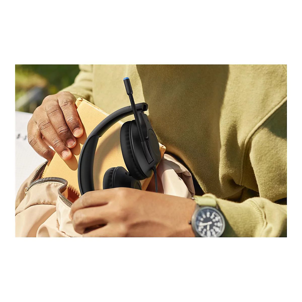 Nahaufnahme: Person hält schwarzes Headset mit Mikrofon. Person trägt grüne Uhr und Pullover. Gelber Notizblock.