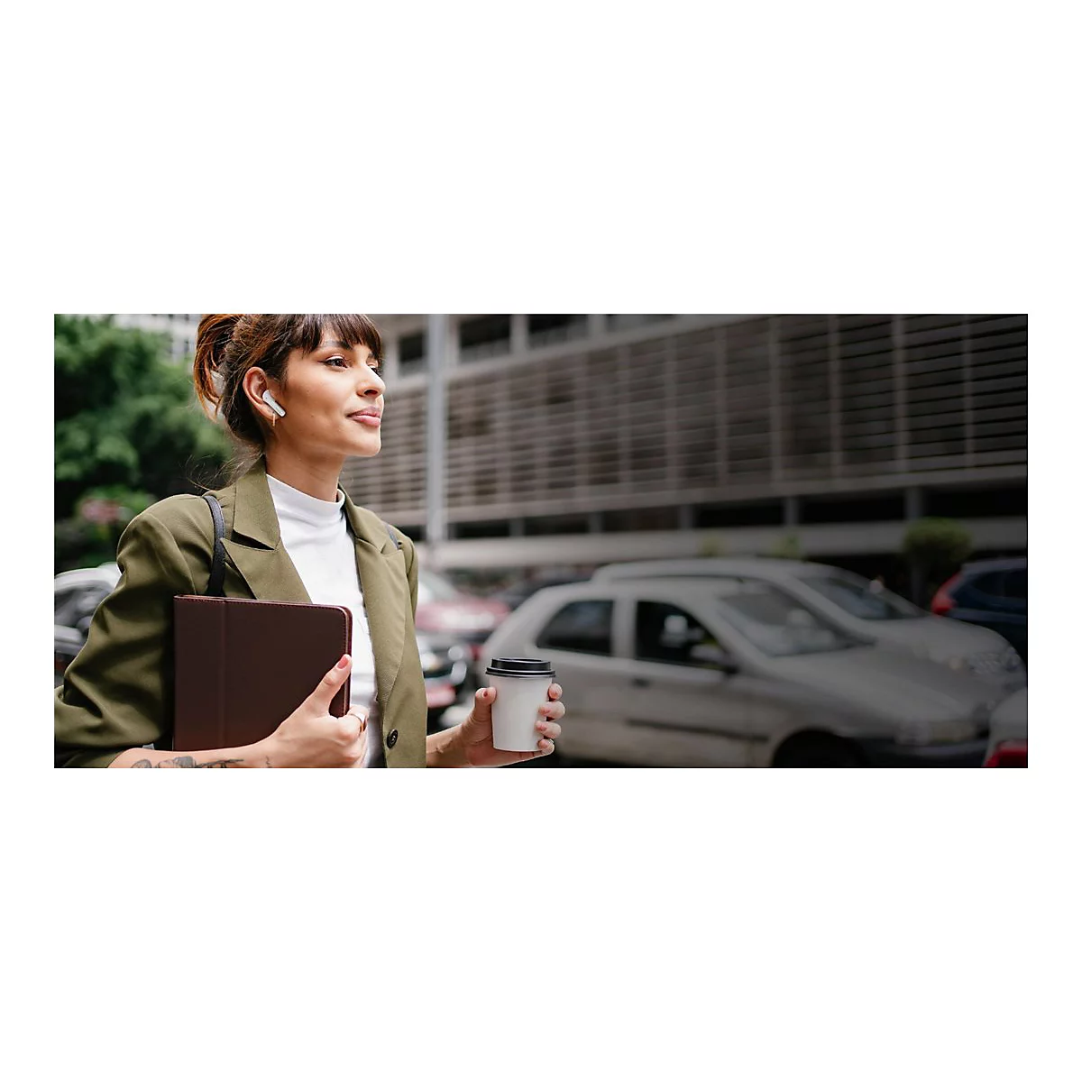 Junge Frau mit Kopfhörern, Tablet und Kaffeebecher; blickt nachdenklich in die Ferne. Sie trägt einen Blazer, steht draußen.