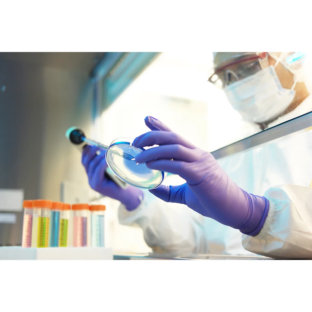 Laborant met veiligheidsbril en handschoenen onderzoekt een petrischaal met een pipet. Reageerbuizen staan op de achtergrond.