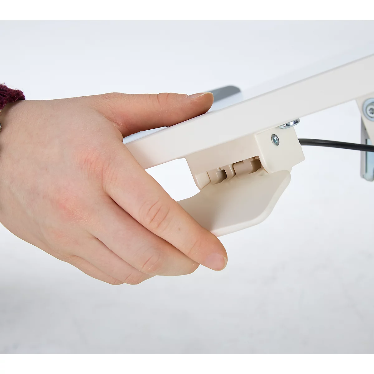 Close-up: Hand die een beige productdetail vasthoudt. Het product is aan een muur gemonteerd. Zwarte kabel, schroeven en gewricht zichtbaar.