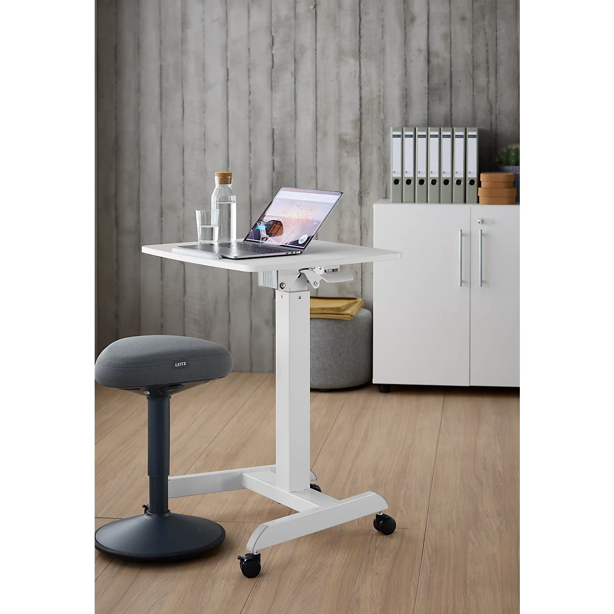Witte, in hoogte verstelbare statafel met laptop, waterfles en bureaustoel. Grijze muur en kast op de achtergrond.