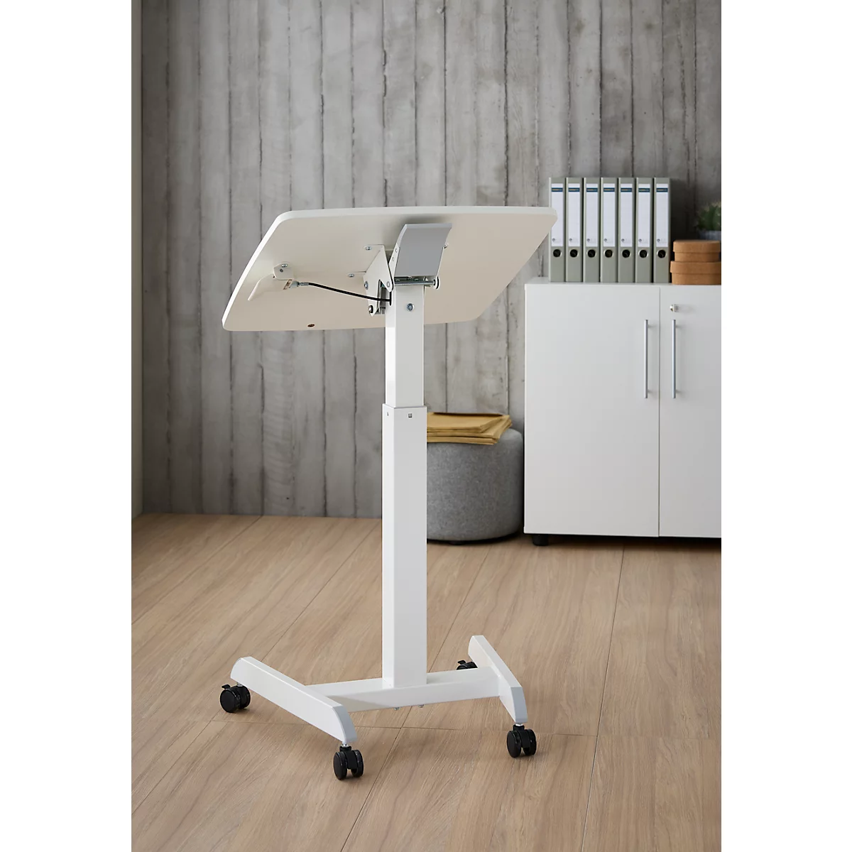 Witte, in hoogte verstelbare statafel met wielen. Tafelblad iets gekanteld, op de achtergrond grijze muur, kast, ordners.