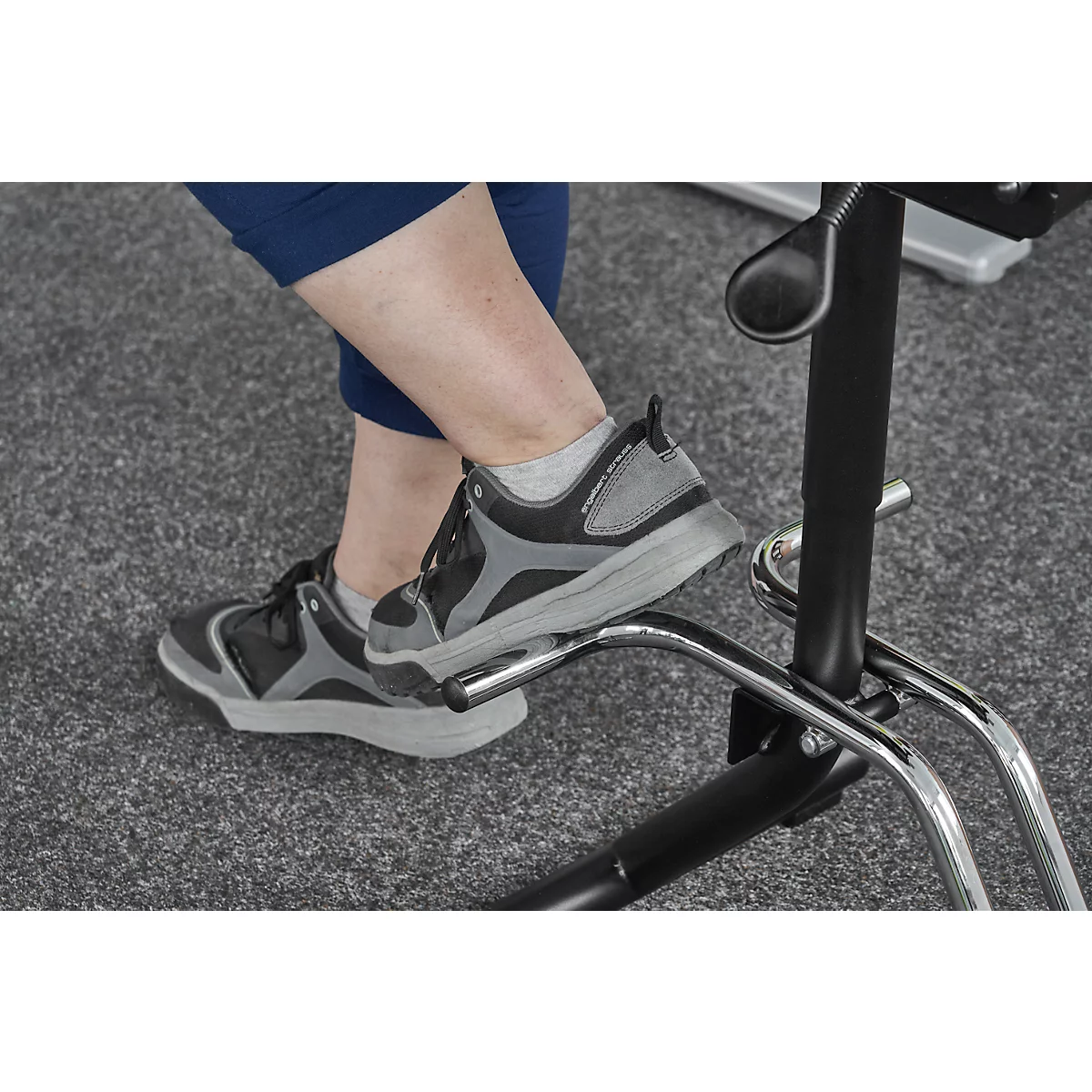 Close-up of feet in gray athletic shoes resting on a footrest of a black chair. The background is a gray carpet.