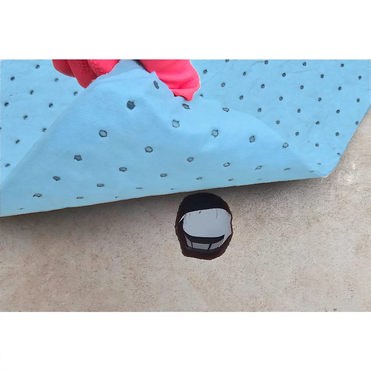 A blue absorbent mat with small dark dots covers a spill. A hand in a pink glove lifts the mat.