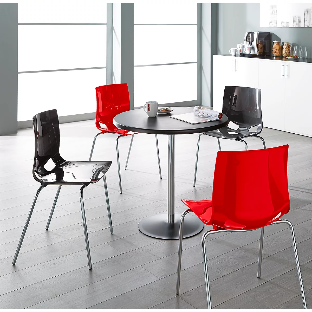 Round table with four chairs, two red, two transparent black. Bright environment.