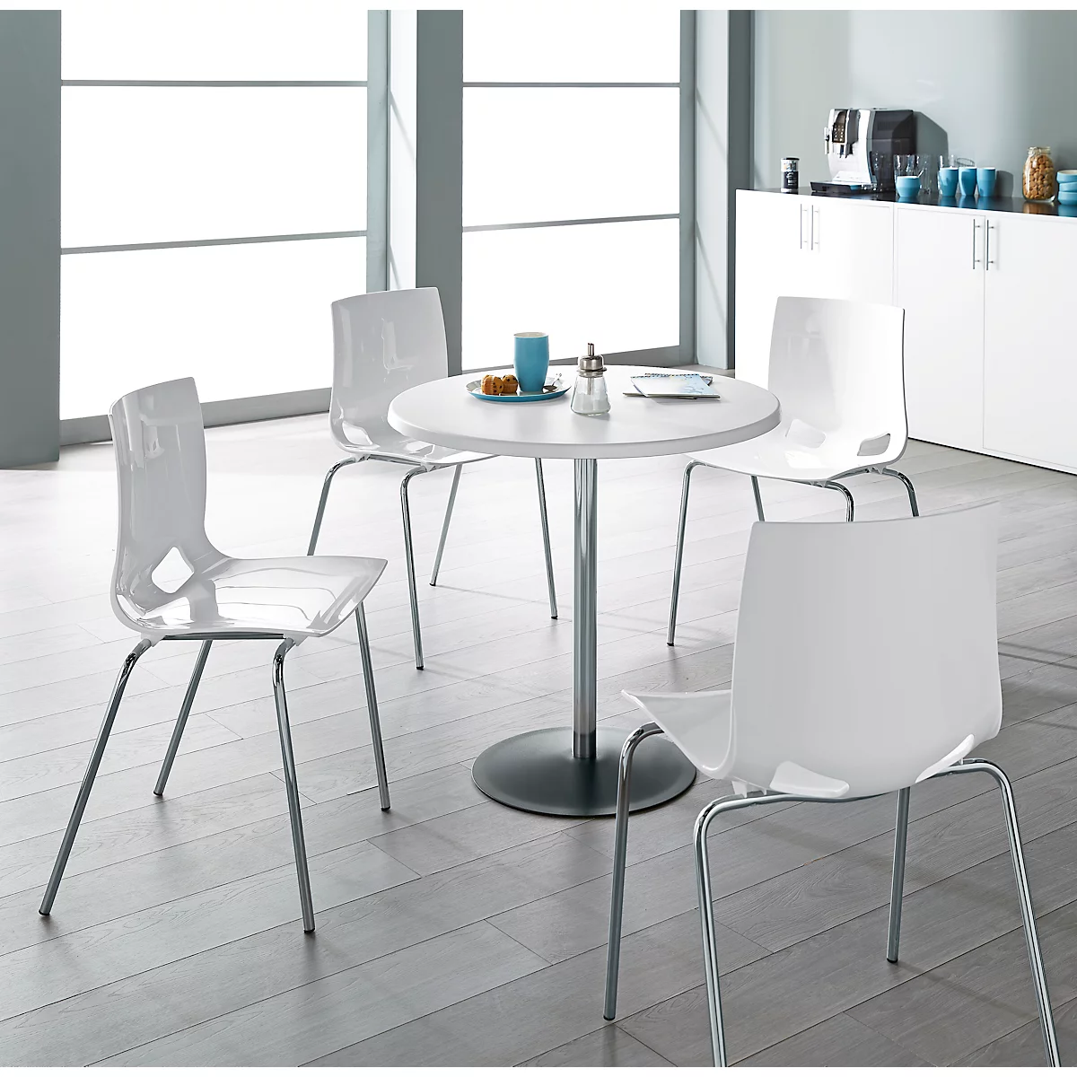 Four white chairs surround a round table. The table has a silver column and a black base. Light wooden parquet.