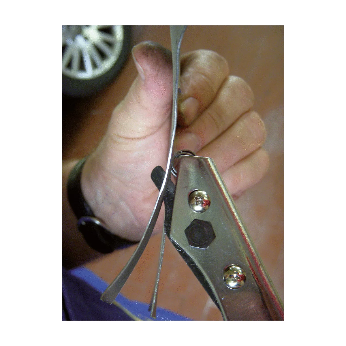 Nahaufnahme einer Hand, die mit einer Lochzange Metall formt. Im Hintergrund ist ein Reifen zu sehen.