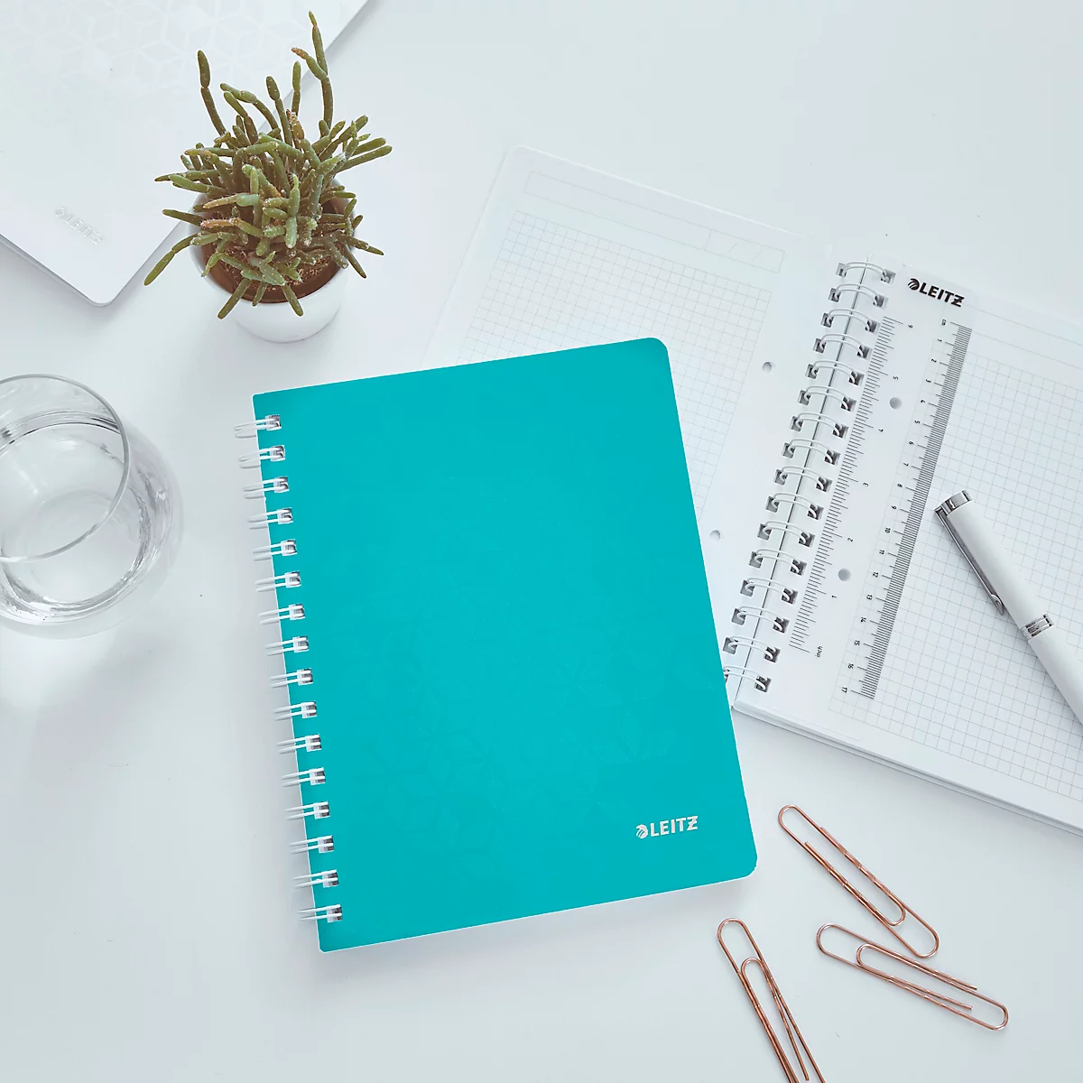 Cahier turquoise avec spirale, à côté règle, stylo et trombones sur surface blanche.