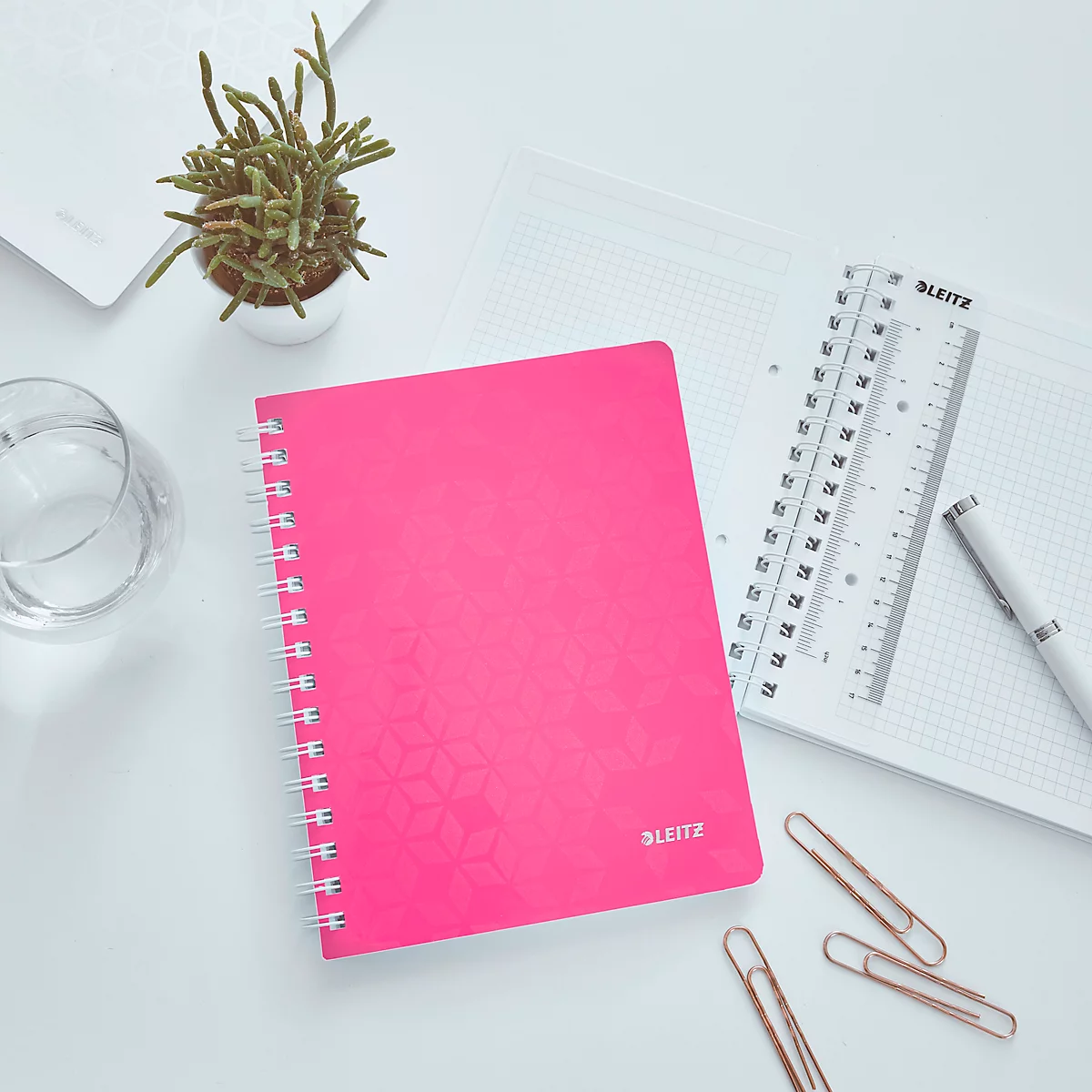 Un cahier rose est posé sur un bureau blanc. À côté, des accessoires, comme un stylo et une règle.