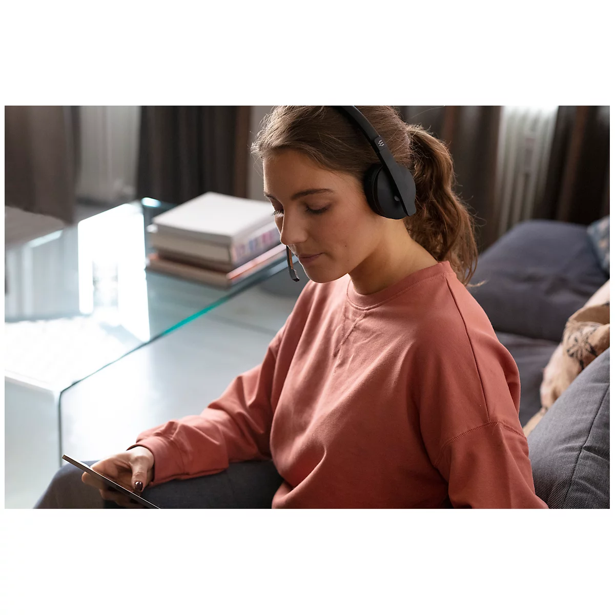 Woman with headphones focused on tablet, sitting on sofa. Background: couch, table and books.
