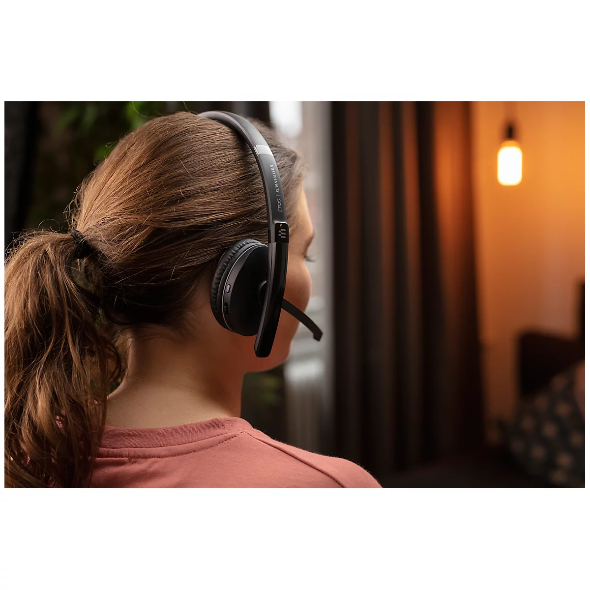 Woman wearing headphones with microphone, looking to the side. Lamp in the background.