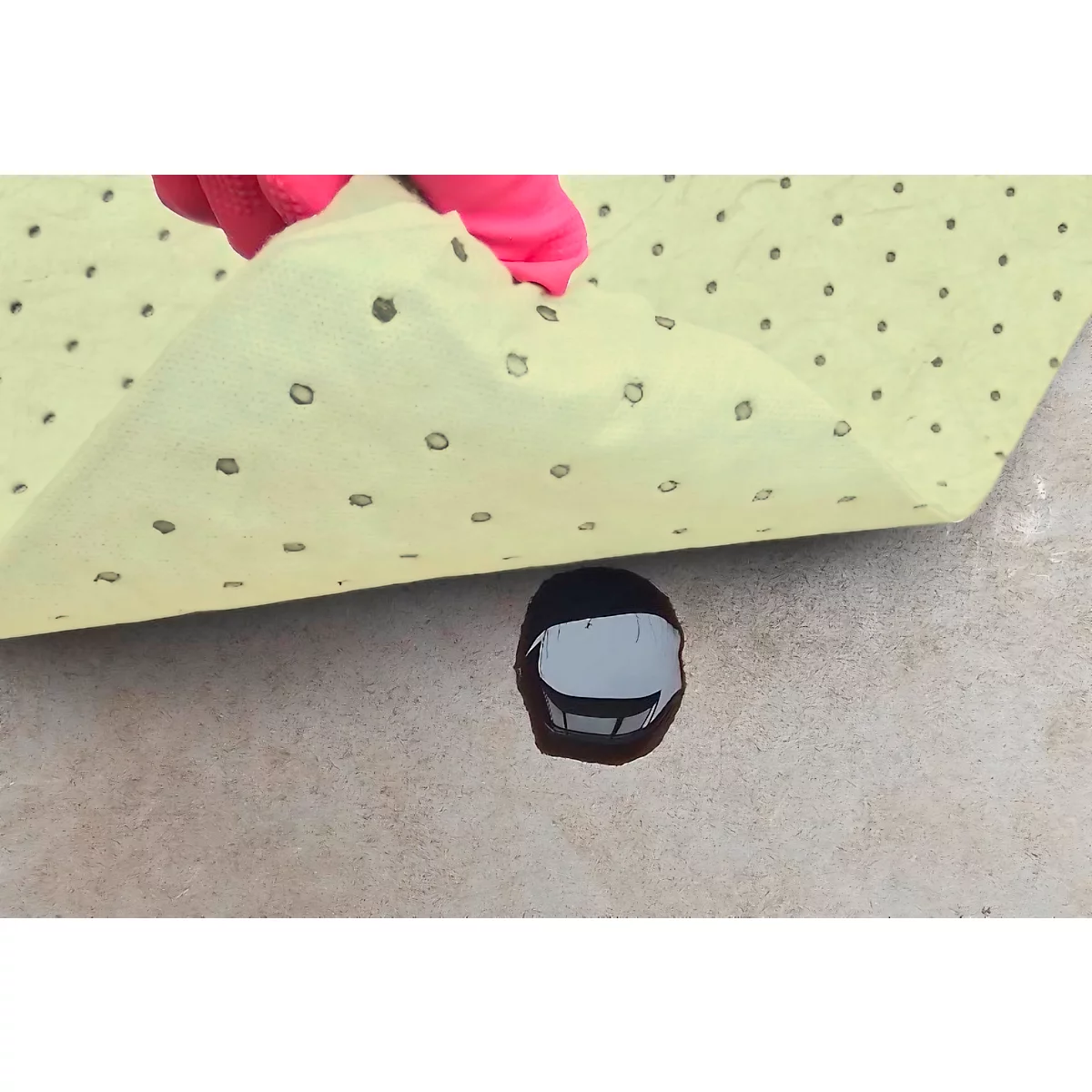 Une main gantée soulève un tampon absorbant l'huile jaune avec des points noirs, révélant un liquide foncé sur un sol gris clair.