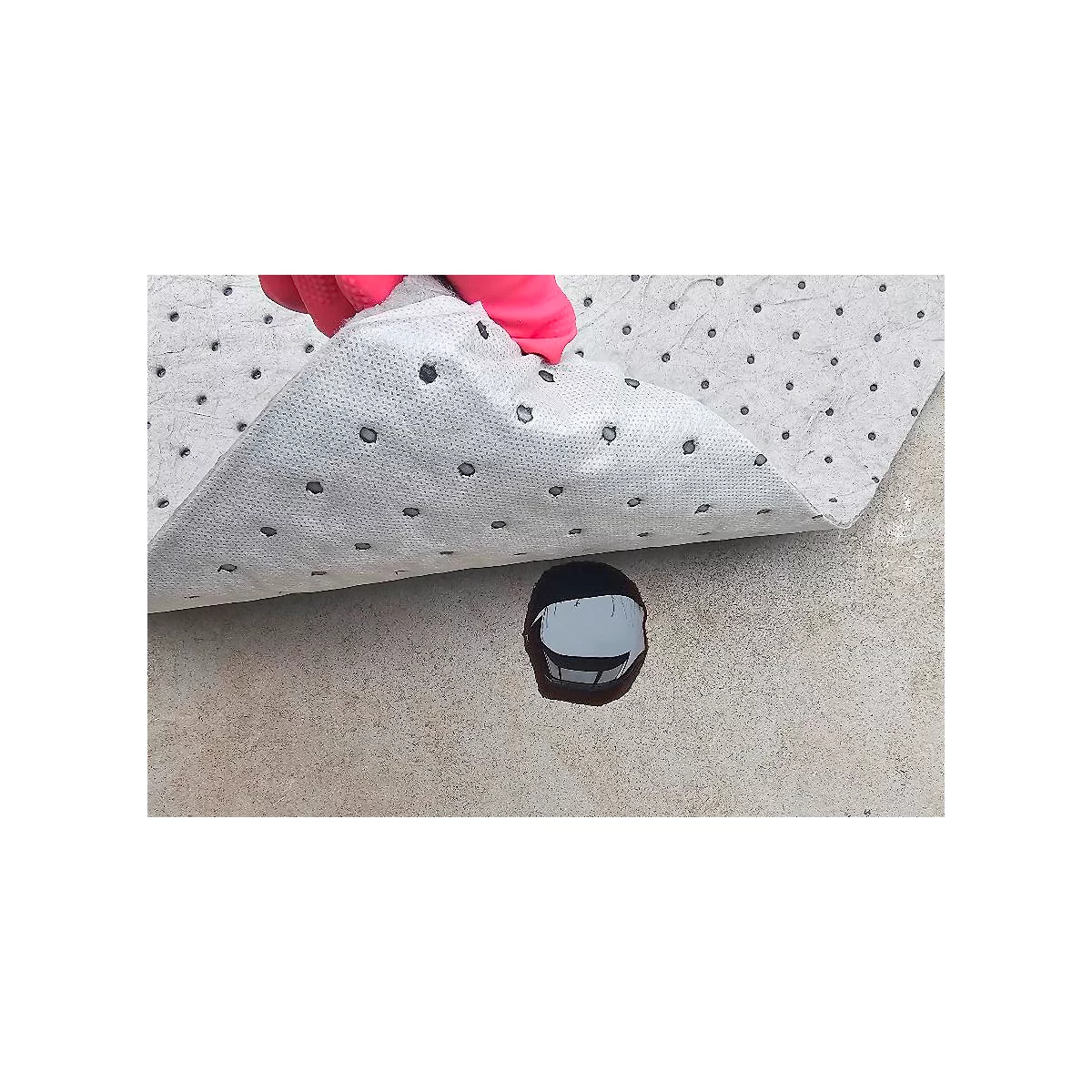 Gant rose soulevant un tapis absorbant l'huile gris avec des points noirs. Liquide sombre en dessous sur une surface claire.