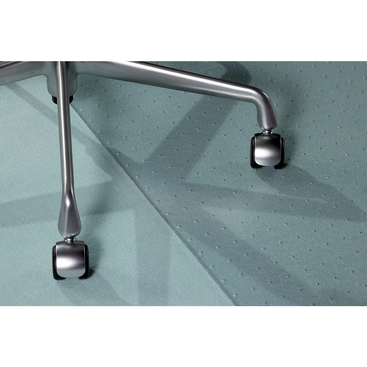 Nahaufnahme von Stuhlrollen aus Metall auf einer strukturierten, hellgrünen Oberfläche. Der Stuhl wirft einen Schatten.