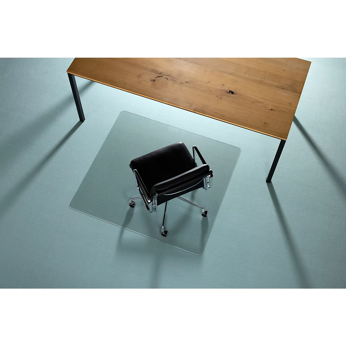 Bürostuhl auf einer transparenten Bodenschutzmatte unter einem Holztisch mit schwarzen Beinen.
