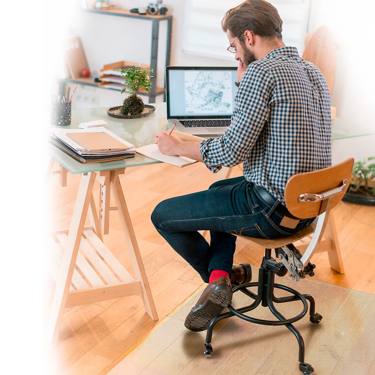 Ein Mann mit Brille sitzt an einem Tisch, er arbeitet am Laptop und schreibt. Er trägt ein kariertes Hemd, Jeans, rote Socken.