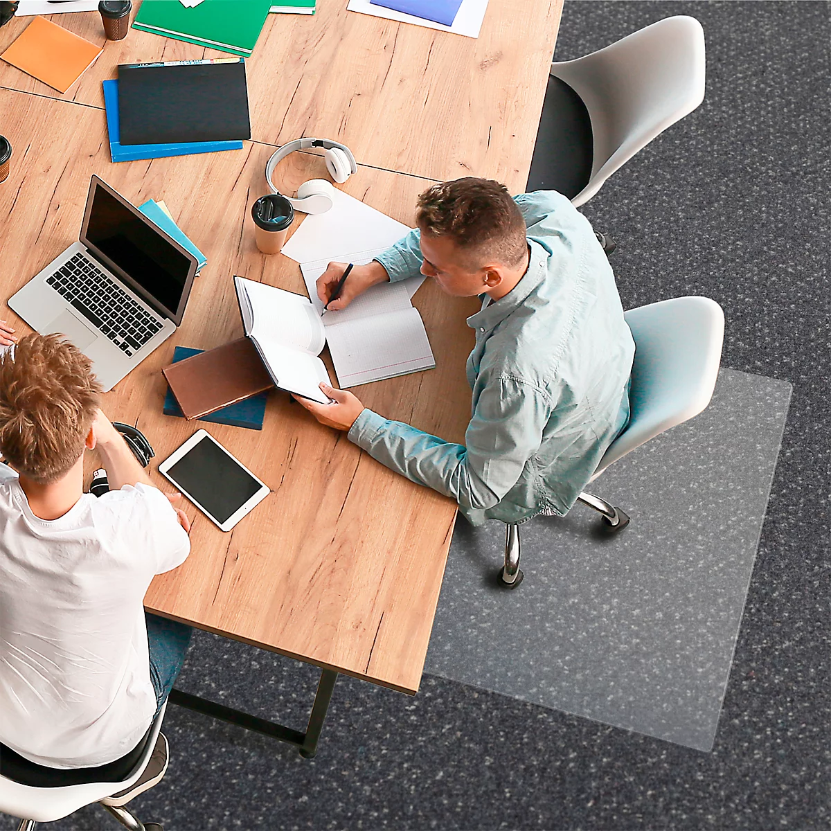 Zwei Personen sitzen an einem Tisch und arbeiten. Eine Person schreibt, die andere blickt auf einen Laptop und ein Tablet.