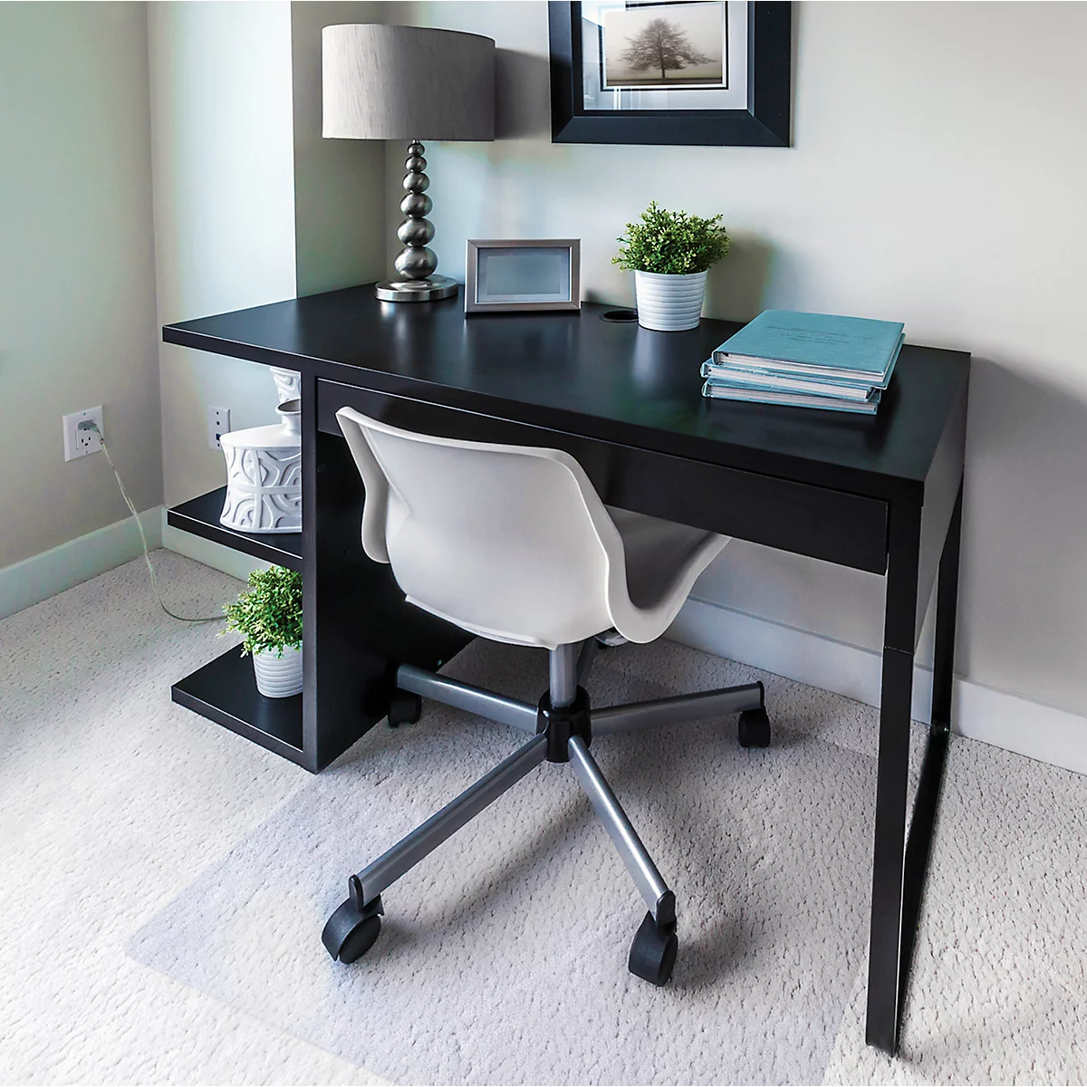 Ein schwarzer Schreibtisch mit weißem Stuhl in einem Arbeitszimmer. Auf dem Tisch stehen Lampe, Pflanzen, Rahmen und Bücher.