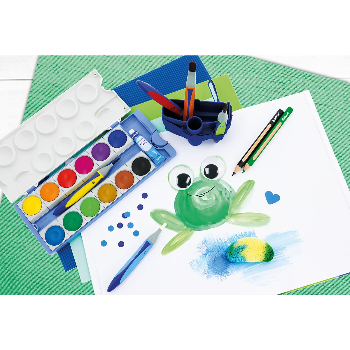 Une boîte de peinture, des crayons et un pinceau sur une feuille de papier représentant une grenouille peinte.