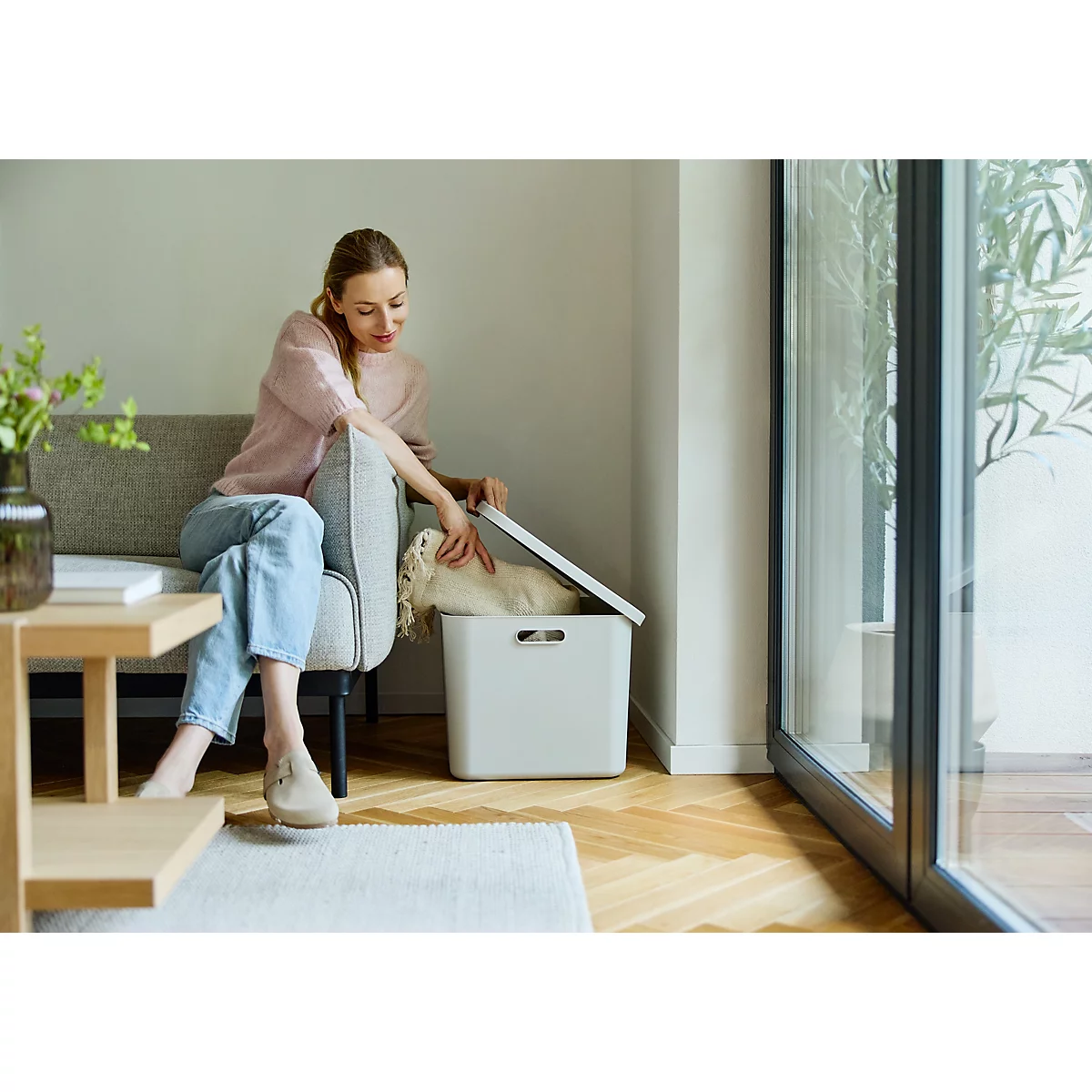 Des boîtes de rangement empilées de différentes tailles et couleurs, à côté d'un canapé. Le sol est en bois, une fenêtre projette des ombres.
