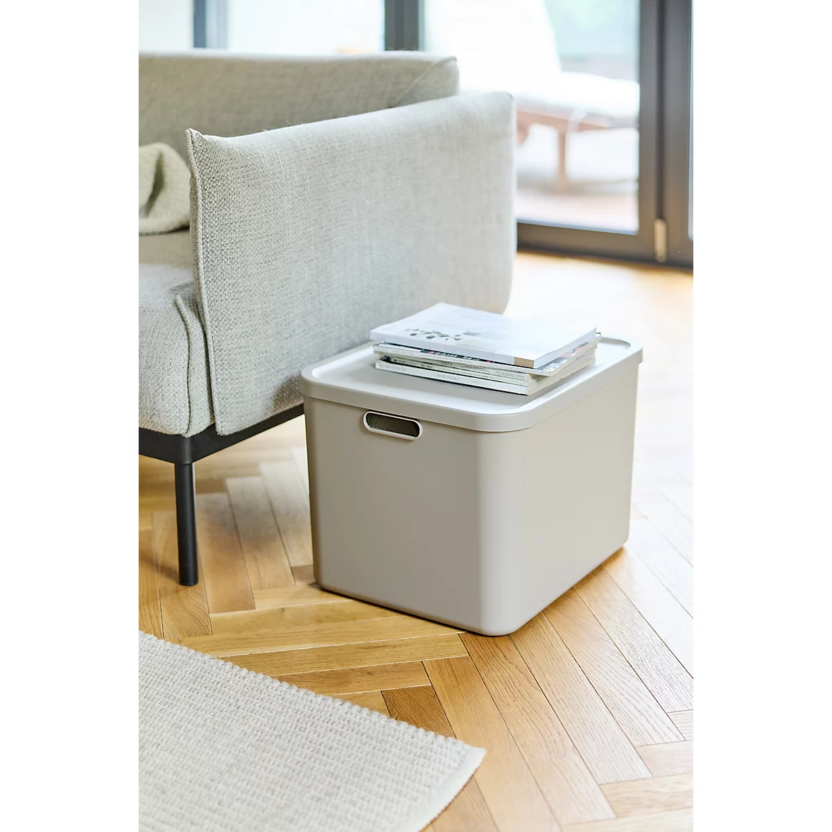 Boîte beige avec poignée. Couvercle avec magazines, à côté d'un canapé gris clair sur plancher en bois.