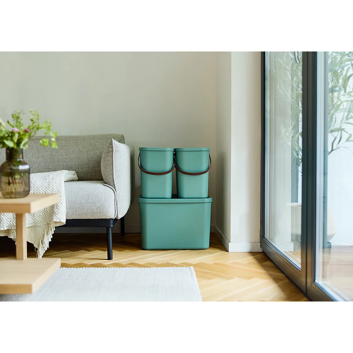 Une femme ouvre une boîte de rangement turquoise sur un canapé, sortant des vêtements. Une table avec des plantes est en arrière-plan.