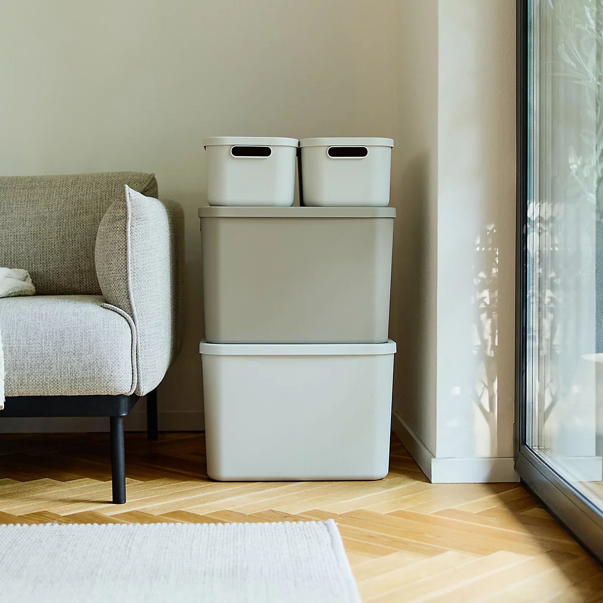 Des seaux et des boîtes gris clair se tiennent devant un canapé clair. Une porte vitrée à droite, une table avec un vase à gauche.