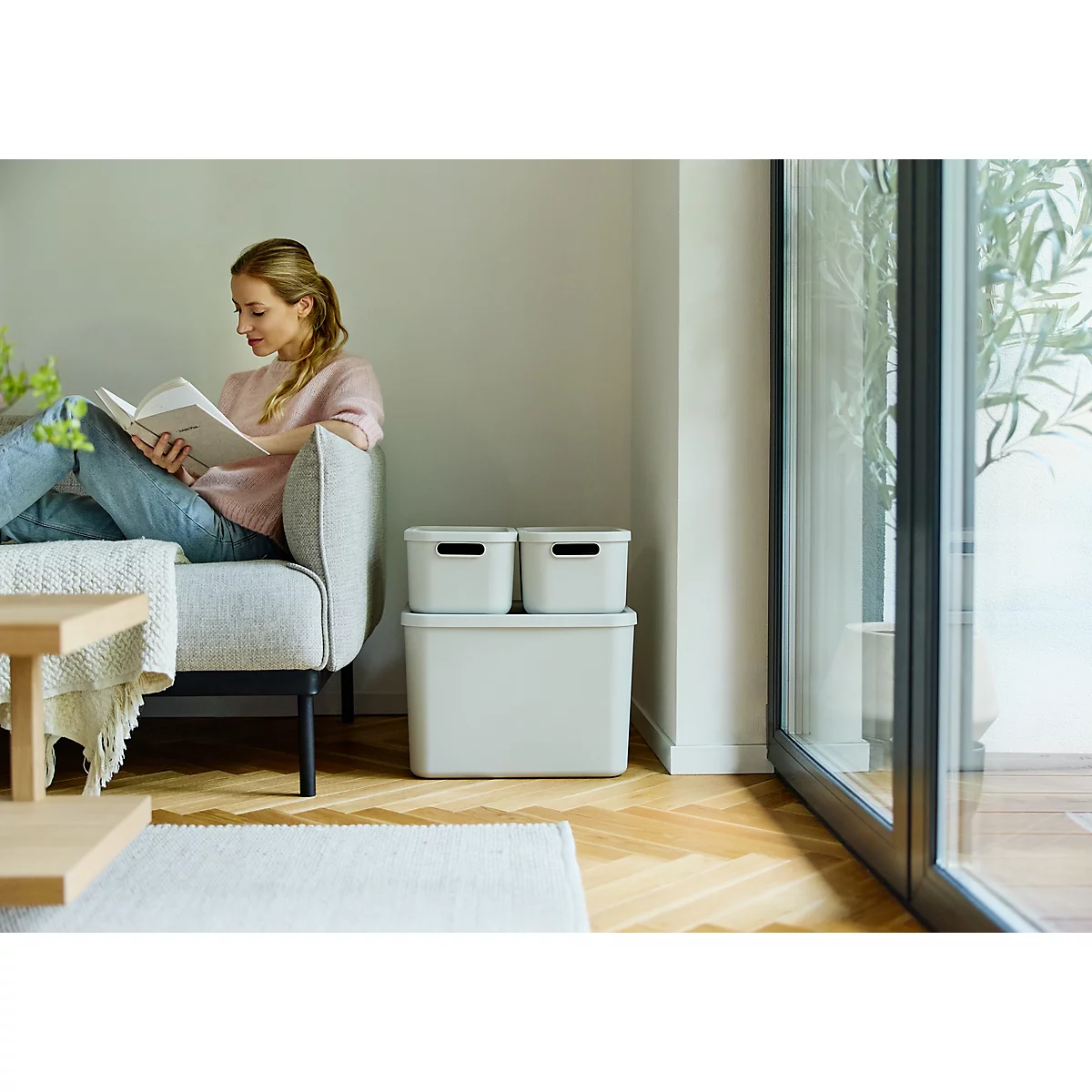 Femme assise sur un canapé lisant un livre, à côté de boîtes de rangement grises.
