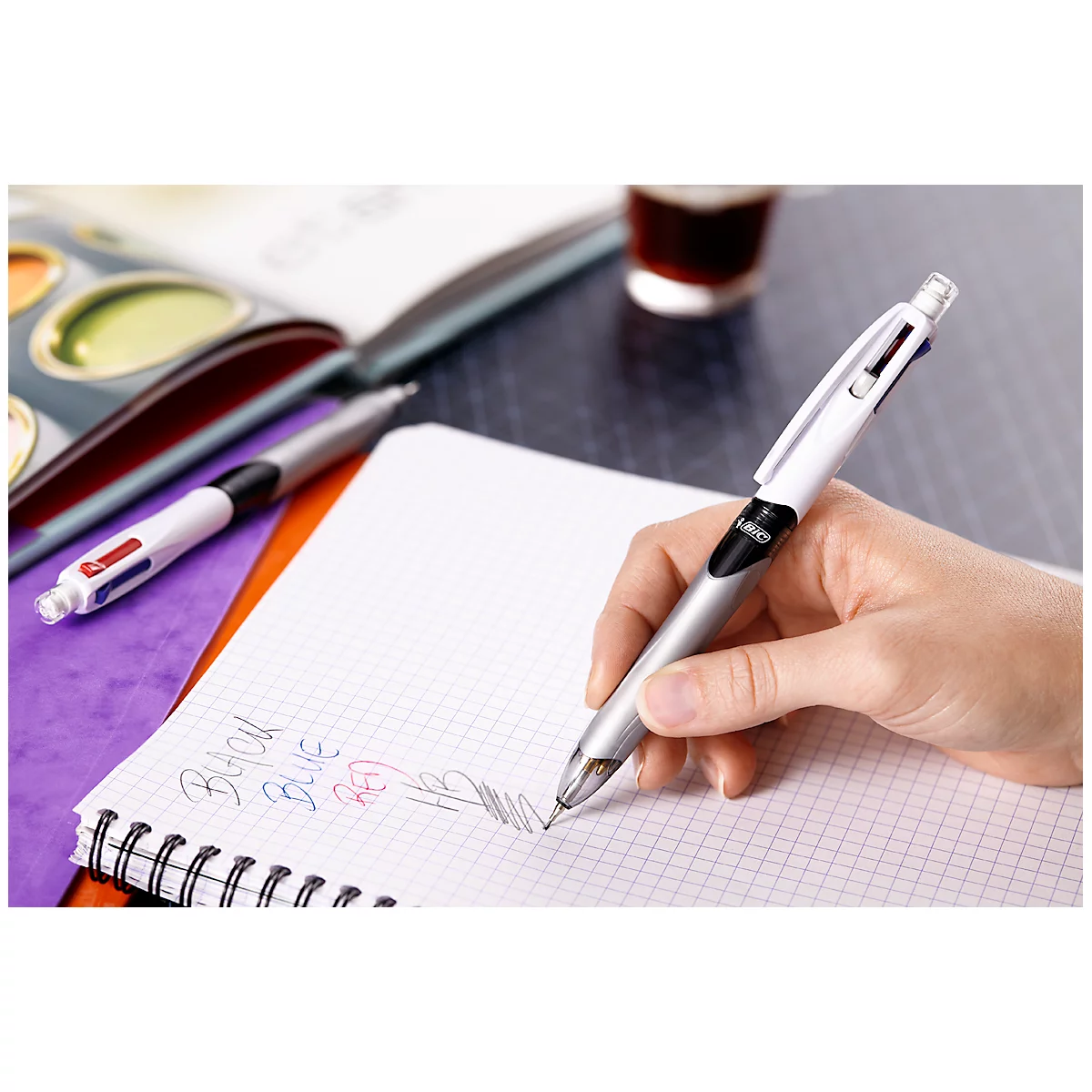 Una mano escribe en un cuaderno cuadriculado con un bolígrafo multicolor blanco.