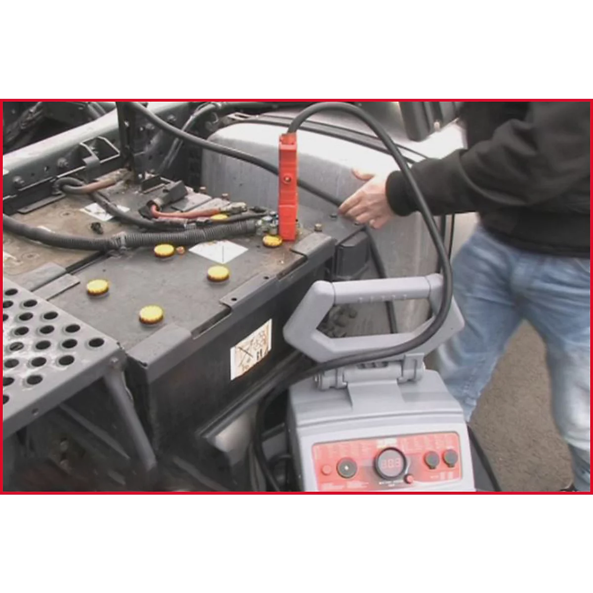 Ein Mann verbindet Starthilfekabel mit der Batterie eines Fahrzeugs. Ein Ladegerät steht am Boden.