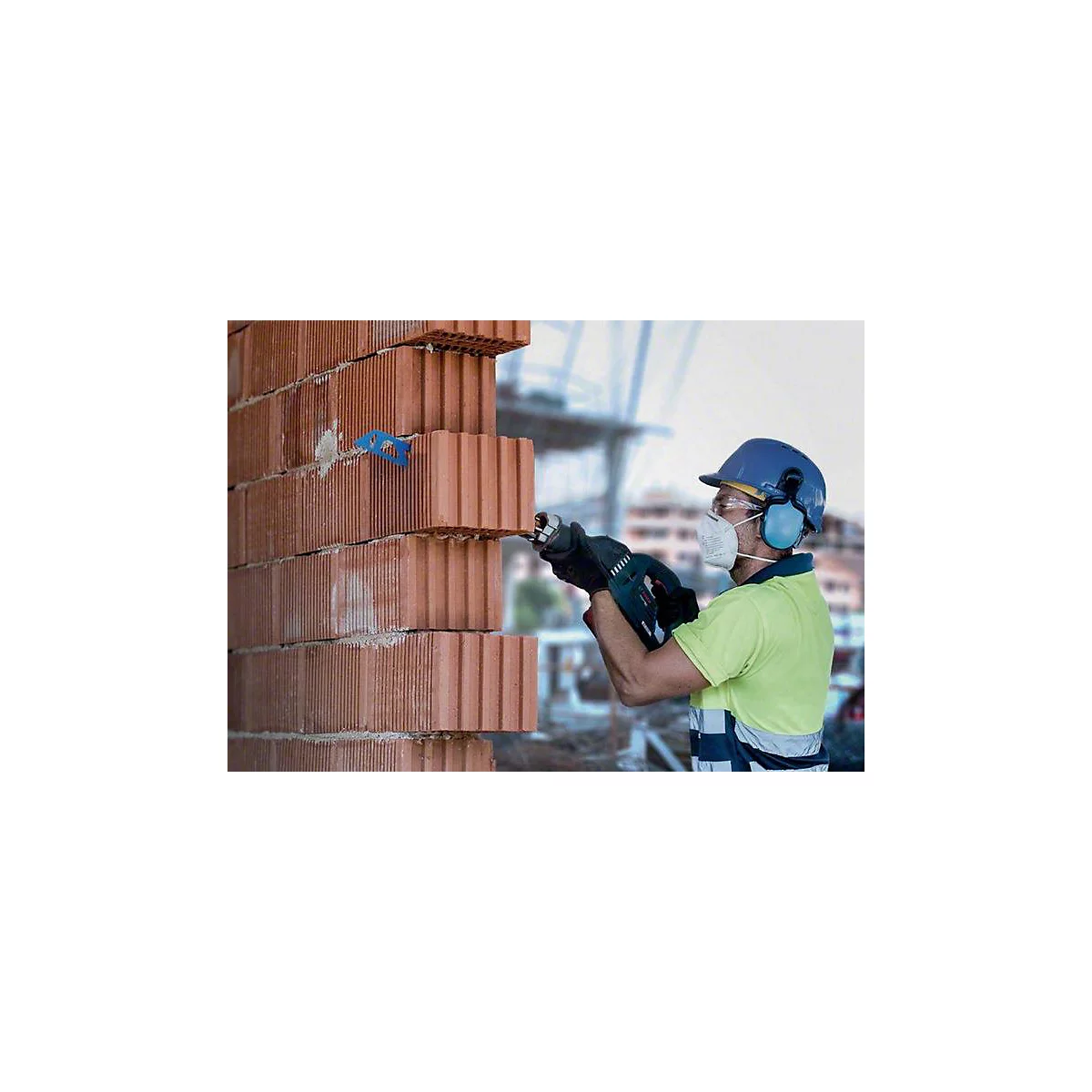 Ein Bauarbeiter mit Schutzhelm und Maske sägt Ziegelsteine. Der Hintergrund zeigt eine Baustelle mit unscharfen Konturen.