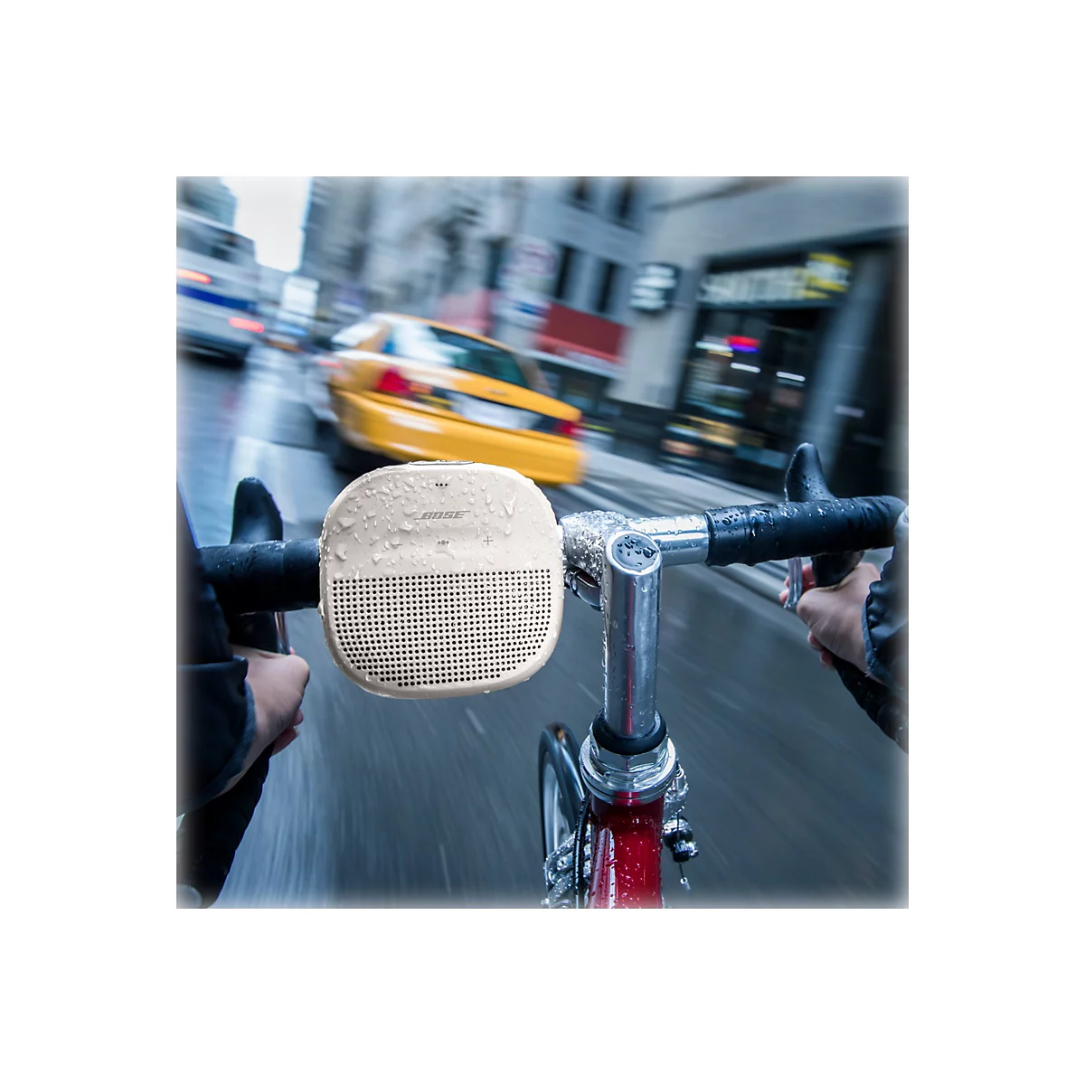 Ein Fahrradfahrer fährt durch die Stadt, ein beigefarbener Bose-Lautsprecher ist am Lenker befestigt. Im Hintergrund verschwommene Bewegung und ein gelbes Taxi.