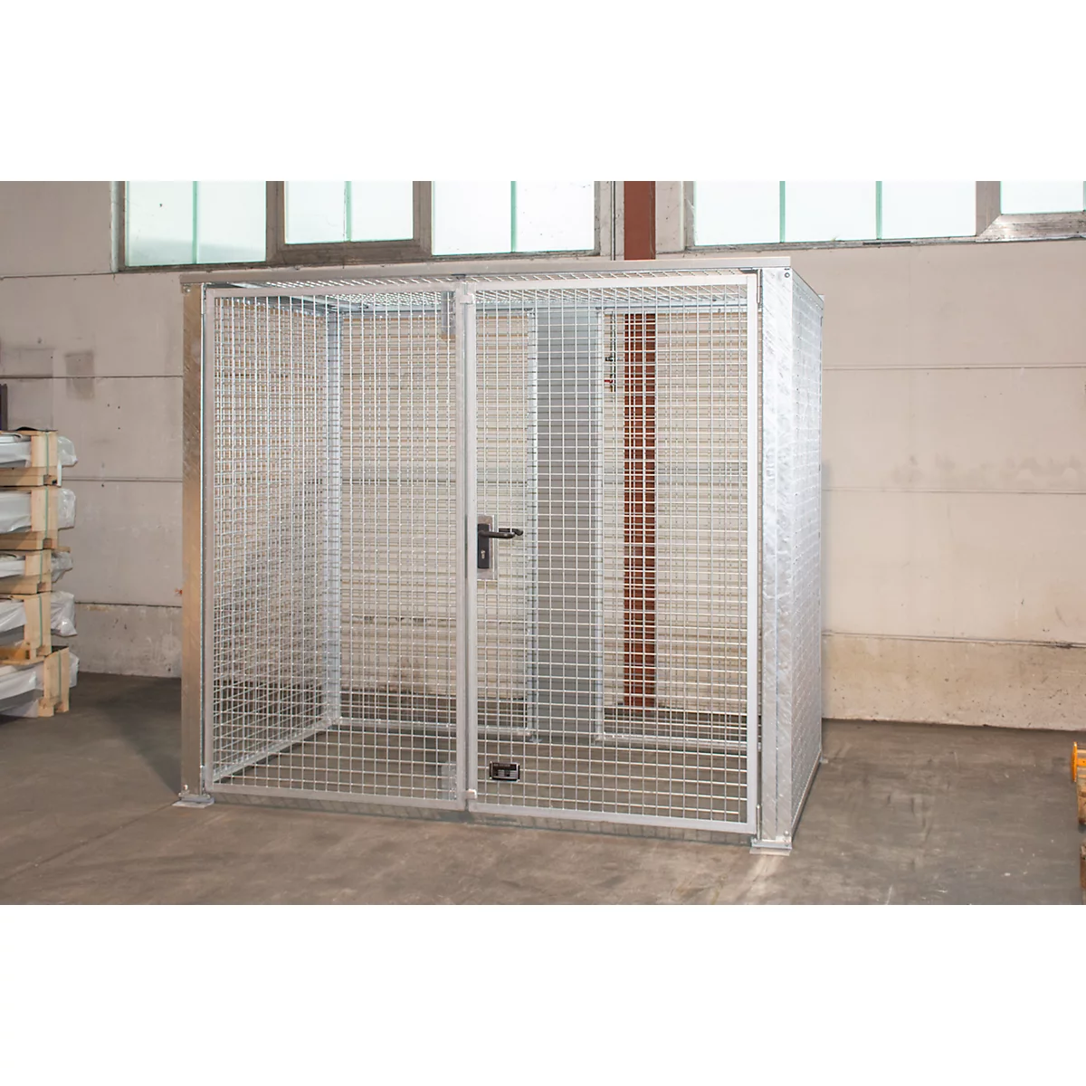 Une cage en treillis métallique argentée avec une porte et une serrure se trouve dans une pièce. Des fenêtres et un mur en béton sont visibles en arrière-plan.