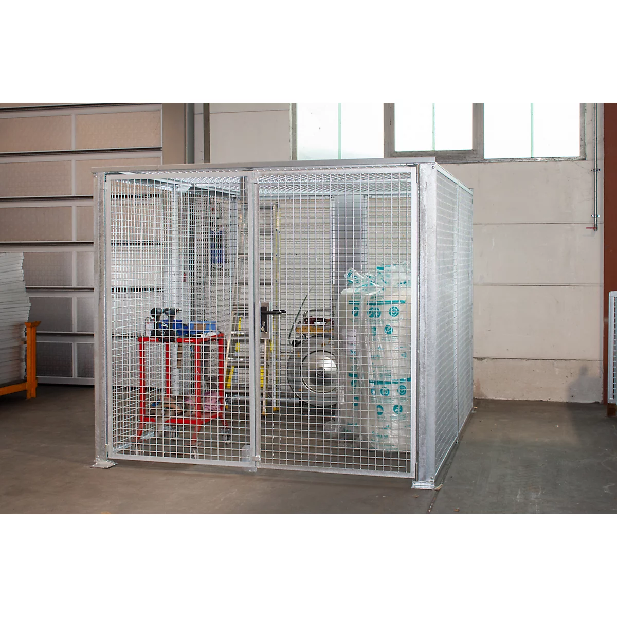 Cage grillagée métallique pour bouteilles de gaz, sur fond clair. À l'intérieur, une table de travail rouge et noire.