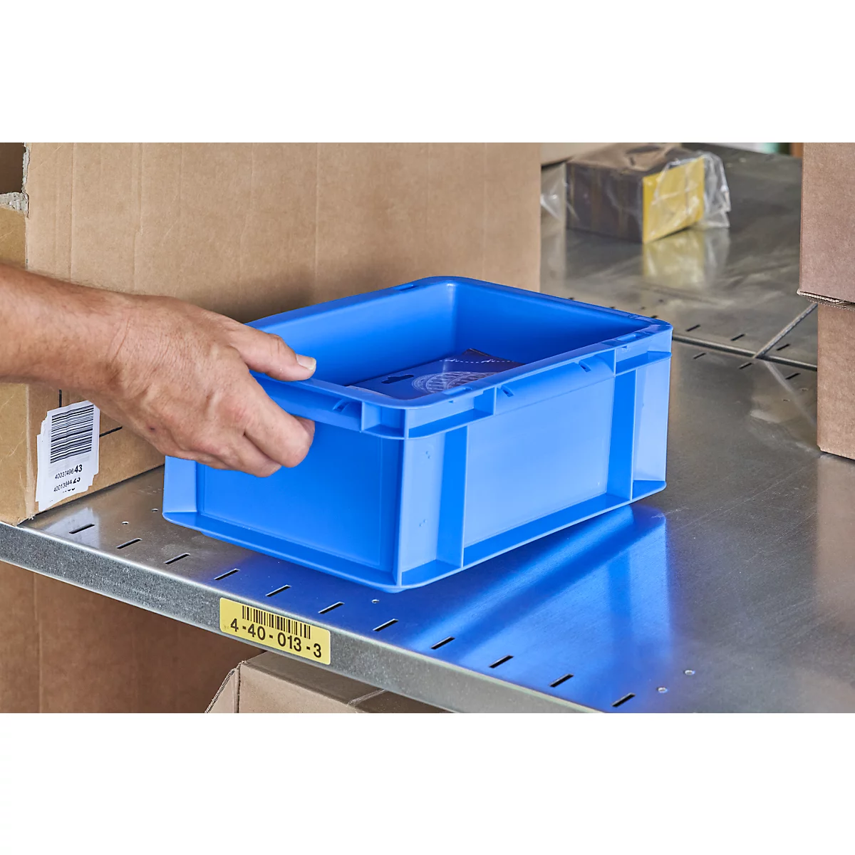 A hand holds a blue plastic box on a shelf. In the background, there are cardboard boxes and other items.