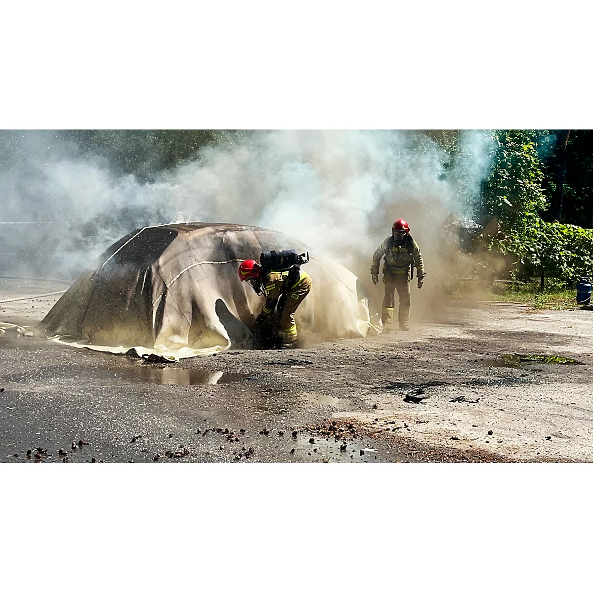 Zwei Feuerwehrleute löschen einen Brand. Ein Feuerwehrmann kniet, der andere steht. Viel Rauch umhüllt einen Gegenstand.