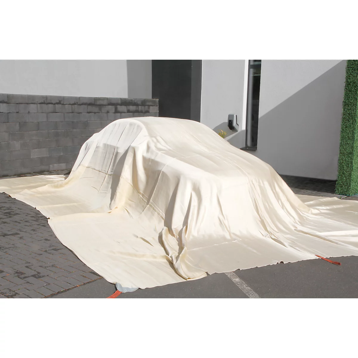 Ein helles Tuch bedeckt ein Auto im Freien. Der Untergrund ist asphaltiert.