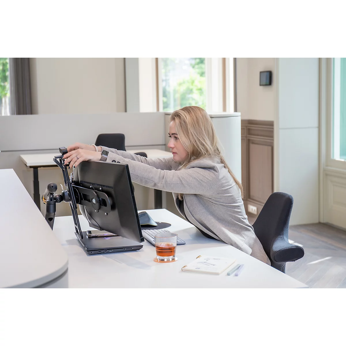 Femme blonde en blazer gris s'étirant au bureau. Elle est assise devant un écran, les mains sur l'écran.