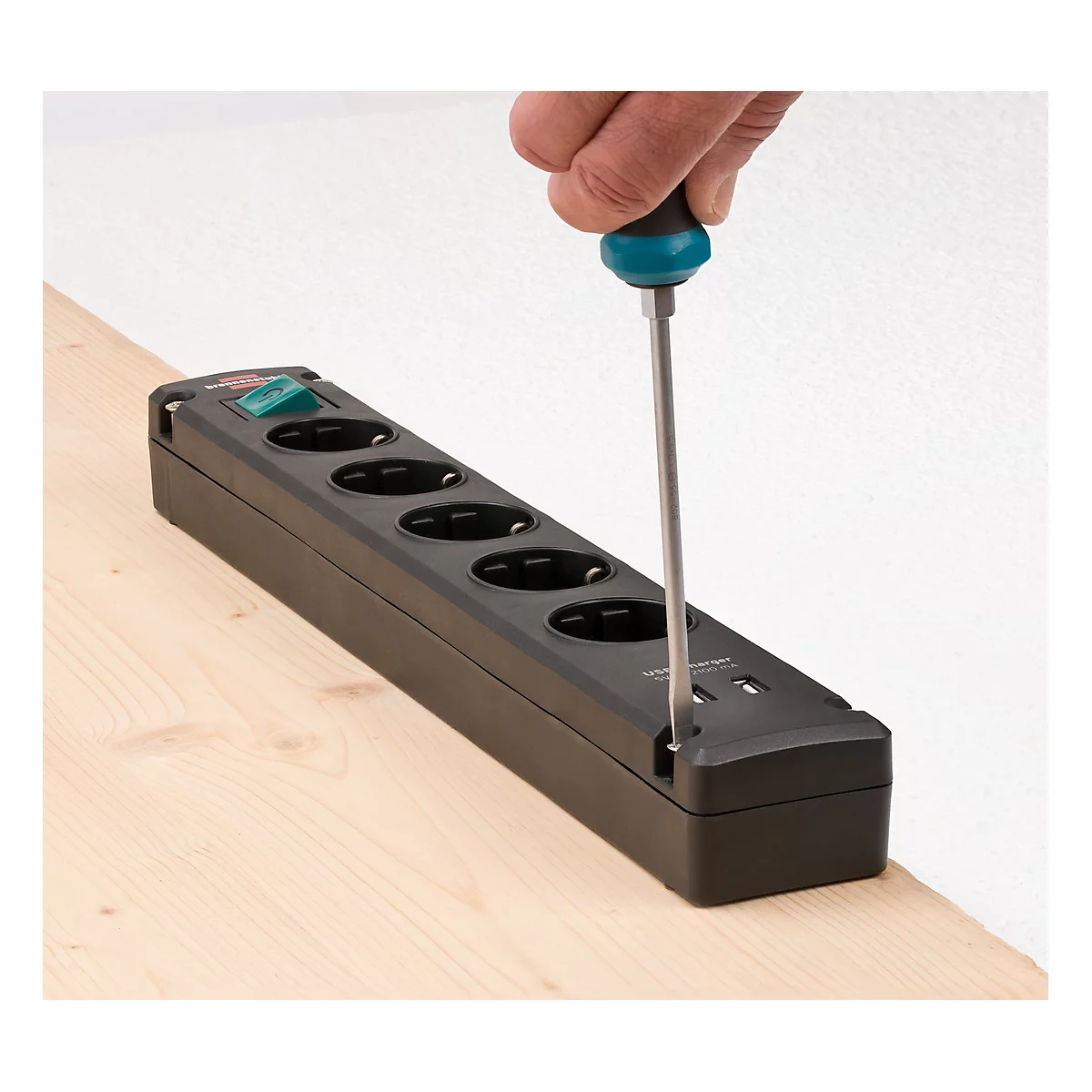 Une personne visse une multiprise noire avec un tournevis, posée sur une surface en bois. À côté, un mur blanc.