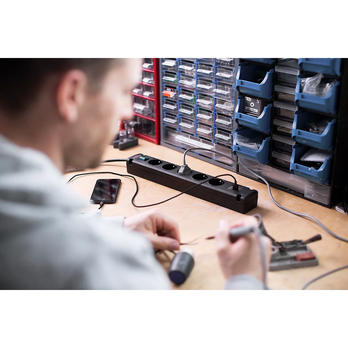 Un homme soude des câbles sur une multiprise. Sur la table, il y a un smartphone, une bobine de soudure et des outils.