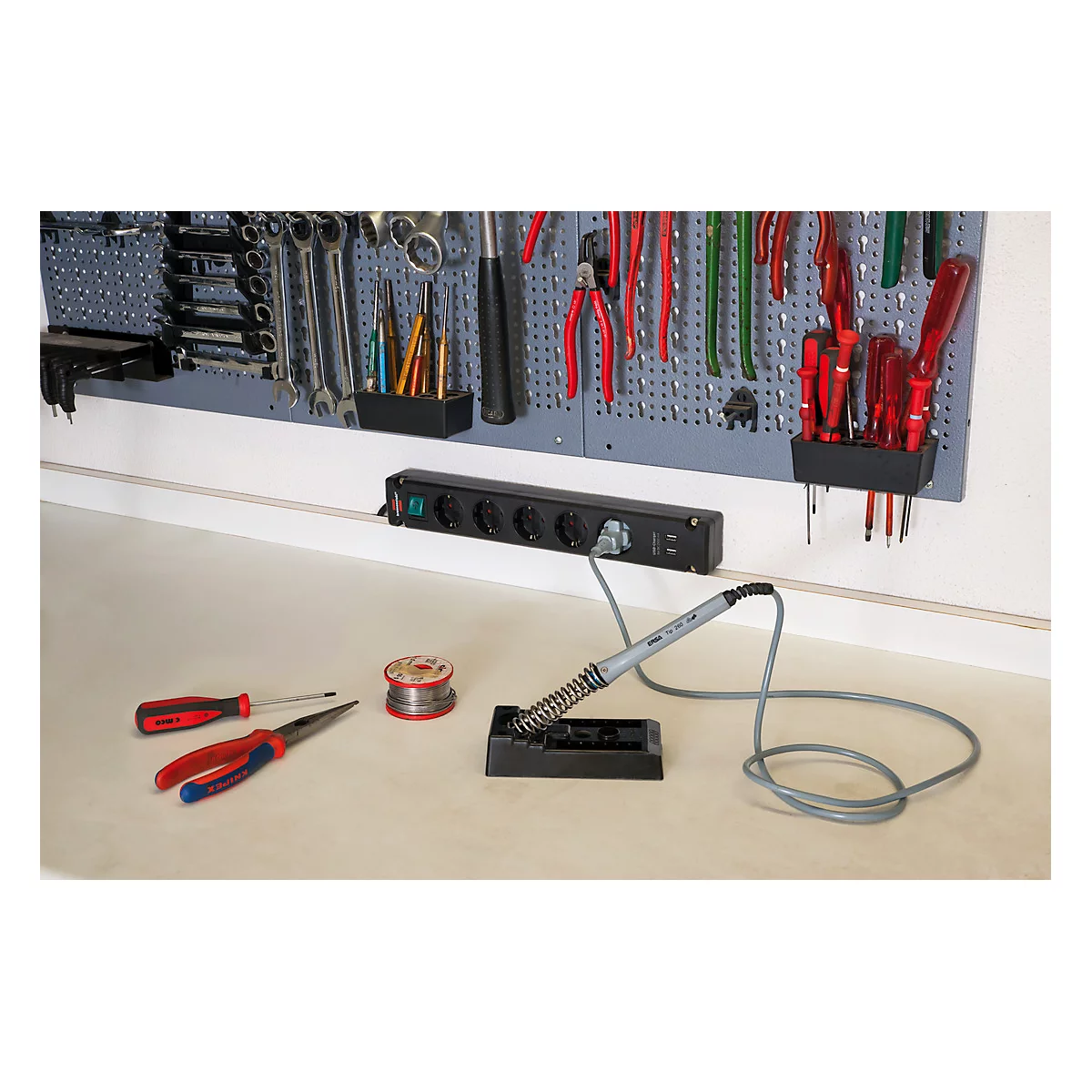 Un établi avec des outils. Une station de soudure est prête, à côté des accessoires et une multiprise.