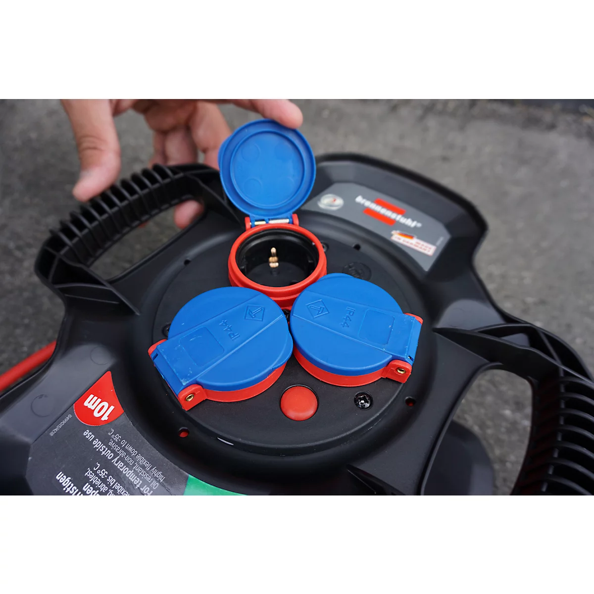 Gros plan d'un enrouleur de câble avec prises ouvertes. Rabat bleu et rouge, bouton rouge, boîtier noir.
