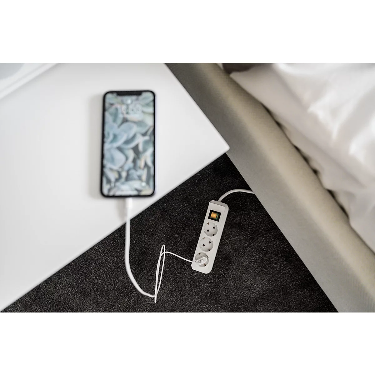 Un smartphone est en charge sur une table de chevet. Le câble blanc relie le téléphone à une multiprise.