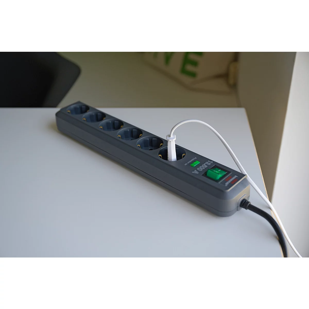 Graue Mehrfachsteckdose auf weißem Tisch. Ein weißes Ladekabel steckt in einer der Buchsen. Ein grüner Schalter ist zu sehen.