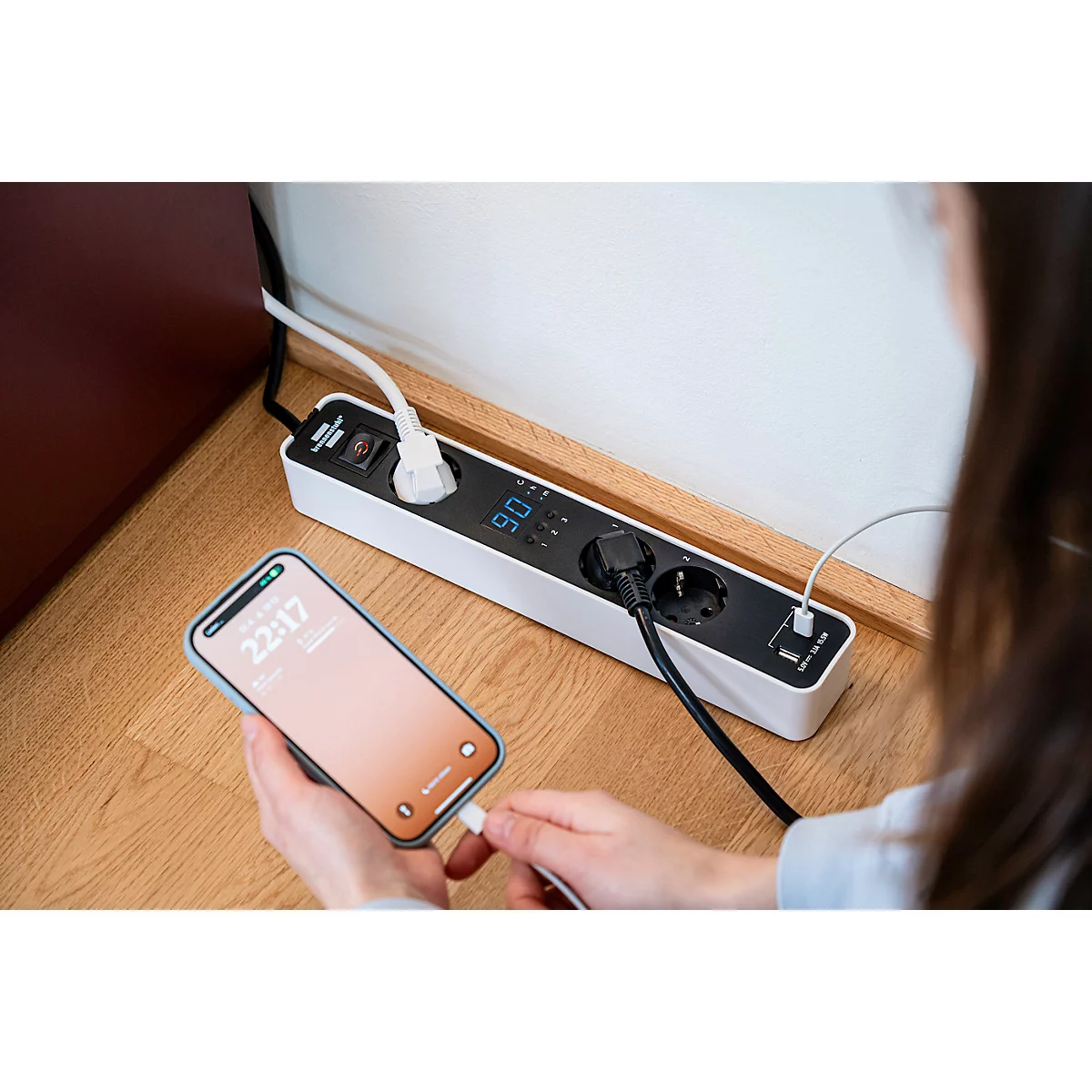 Een witte stekkerdoos met een digitaal display ligt op een houten vloer. Een witte kabel is aangesloten.