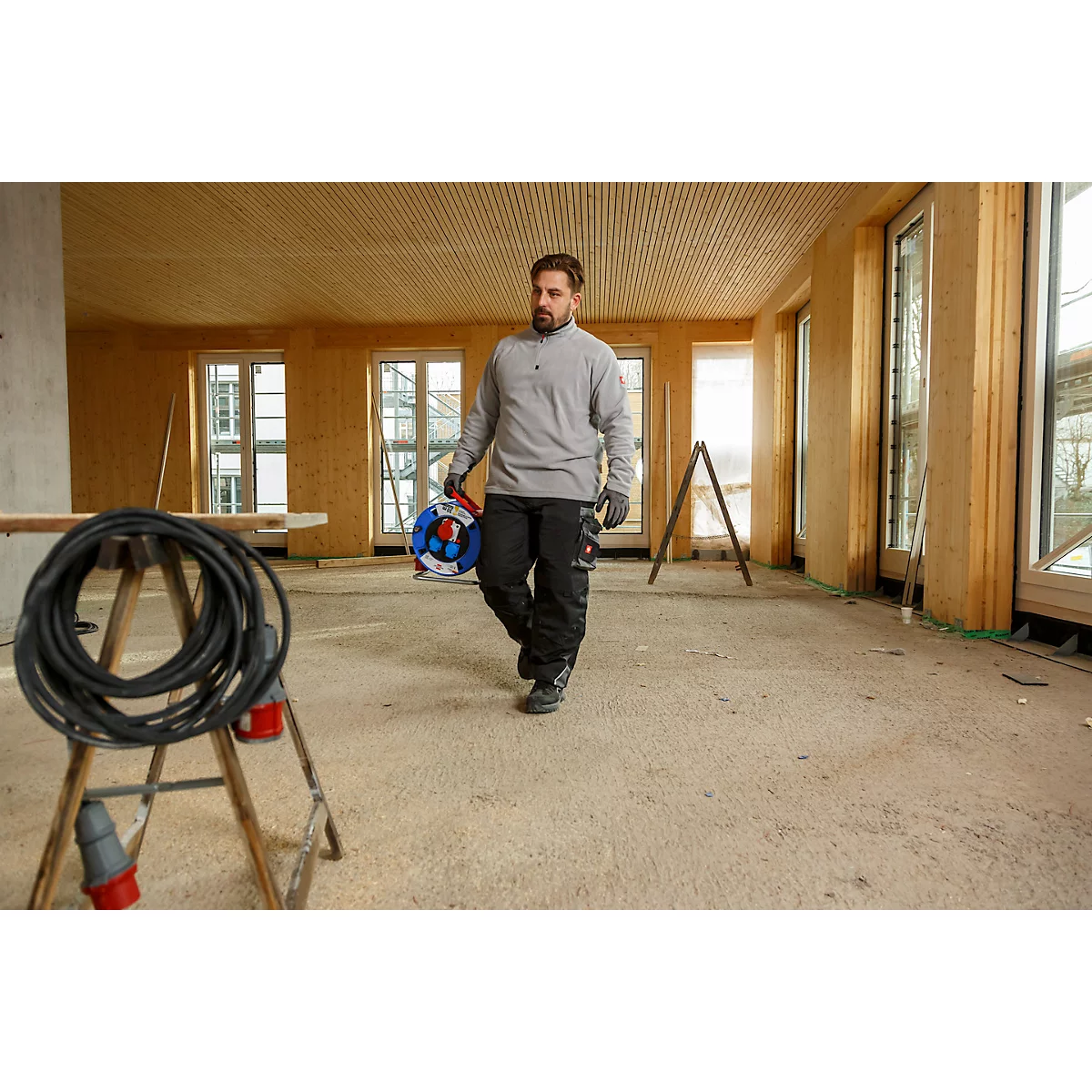 Een man loopt door een kamer met houten bekleding, met een kabelhaspel in zijn hand. Op de voorgrond een kabel, links een tafel.